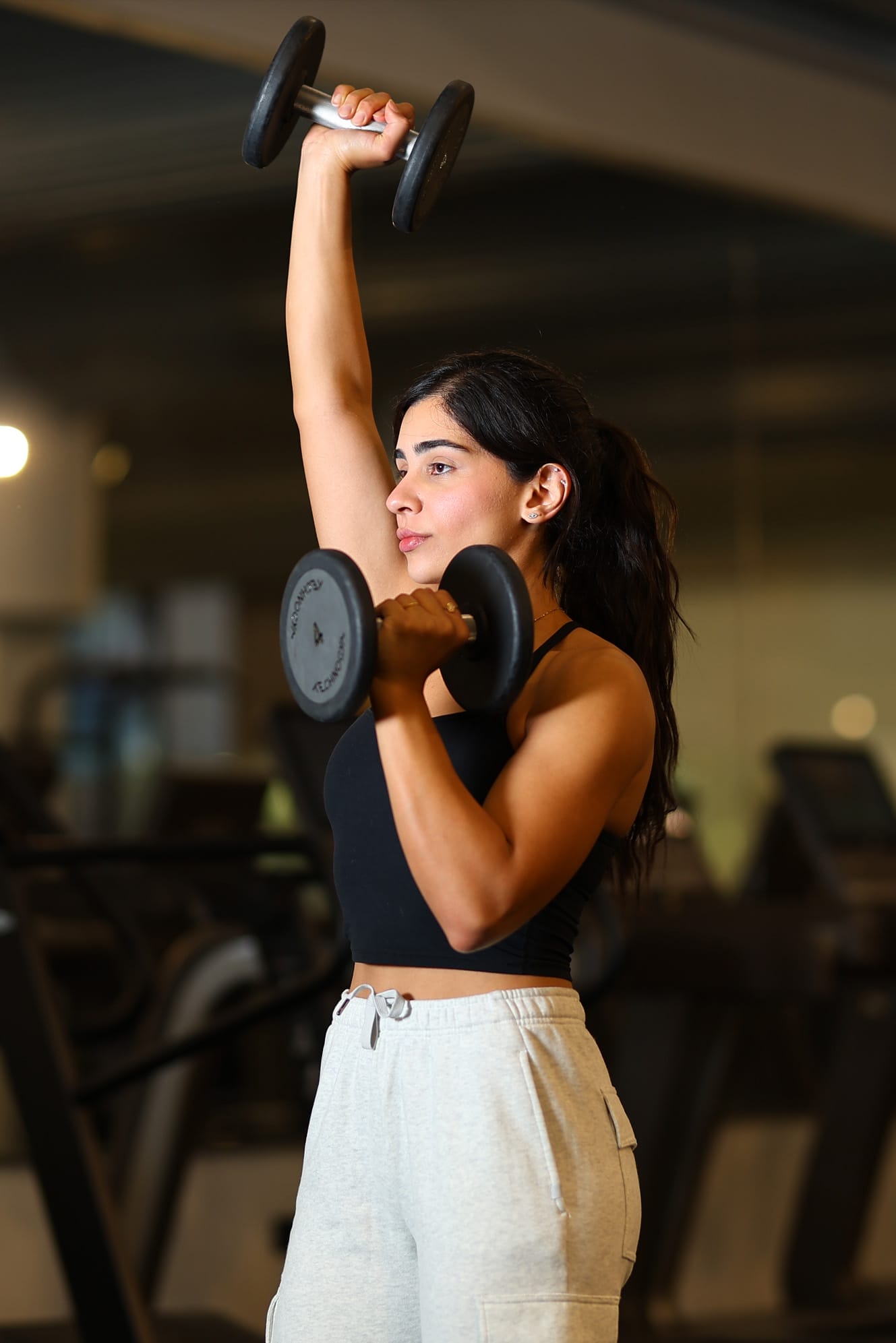 A woman in a black tank top and light gray sweatpants lifts two dumbbells at the gym, holding one weight above her head and the other at shoulder height. She has long dark hair pulled back and appears focused on her workout. Gym equipment is blurred in the background.
