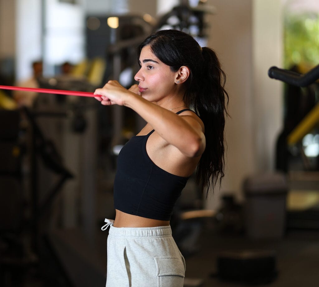 A woman wearing a black crop top and gray sweatpants exercises in a gym, pulling a red resistance band horizontally at chest level. She has long dark hair tied back and focuses ahead; gym equipment is blurred in the background.