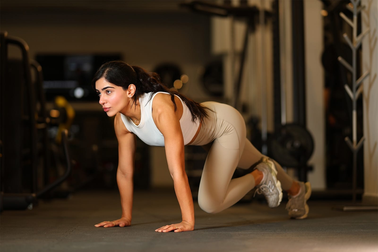 A woman in light workout clothes and sneakers performs a mountain climber exercise on a gym floor. She is focused, with her hands on the ground and knees alternately driving forward, surrounded by gym equipment in the background.