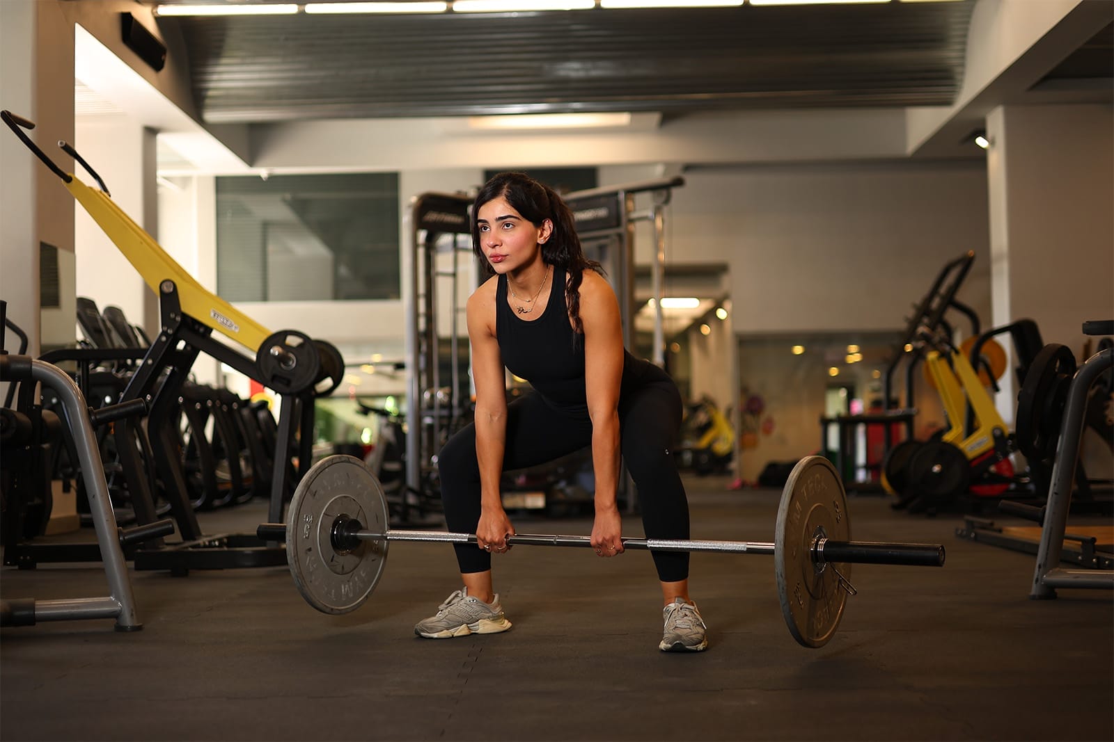 A woman in black athletic wear and gray sneakers prepares to lift a barbell in a gym. She is in a wide stance with bent knees, gripping the bar on the floor, surrounded by gym equipment in a well-lit, modern fitness facility.